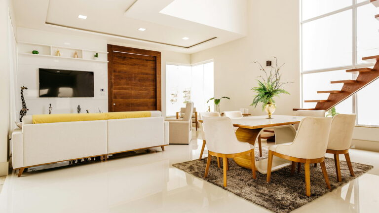 A sophisticated modern interior featuring a cream sofa, sleek dining set, and floating staircase bathed in sunlight — blending minimalism with luxury warmth.