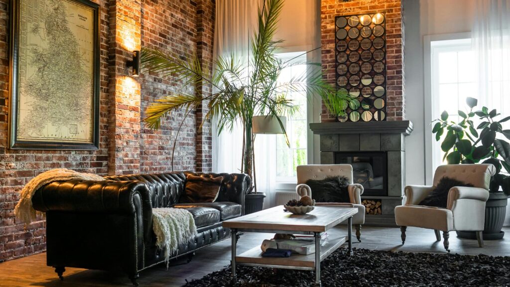 A sophisticated industrial-style living room featuring a black Chesterfield sofa, brick walls, vintage map art, and a cozy fireplace surrounded by greenery — blending rustic charm with modern elegance.